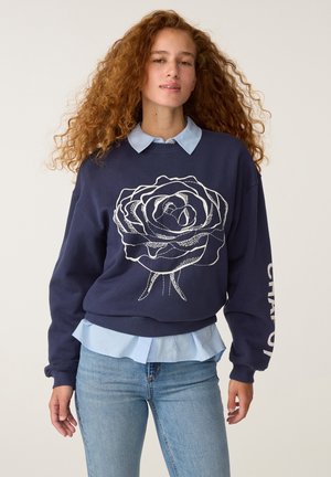 Jonge vrouw met krullend rood haar die een marineblauwe trui draagt met een groot wit rozenpatroon en een lichtblauw overhemd eronder.