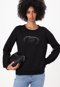 Sudadera negra de manga larga, con cuello redondo y un diseño gráfico texturizado en el frente. Modelo sosteniendo un pequeño bolso negro de mano.