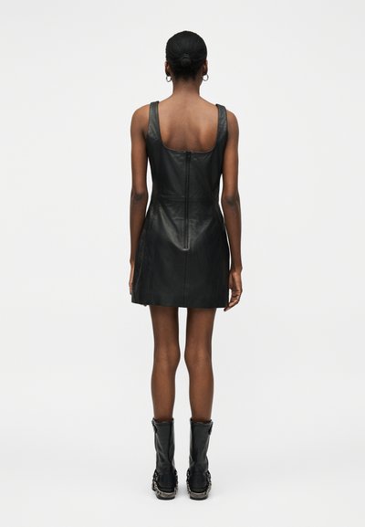 Γυναίκα με σκούρο δέρμα και μαλλιά σε κότσο, που φοράει ένα μαύρο δερμάτινο μίνι φόρεμα χωρίς μανίκια και μαύρες μπότες μέχρι τη μέση της γάμπας, στέκεται με την πλάτη στραμμένη.