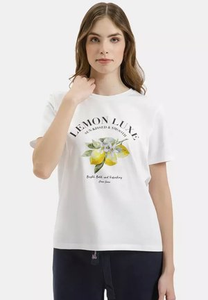 Młoda kobieta w białej koszulce z nadrukiem cytryny i napisem "LEMON LUXE sun-kissed & smooth" na jednolitym tle.