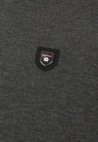 Dunkelgrauer Strickpullover mit einem kleinen schwarzen Wappen-Patch mit metallischen Akzenten, auf der linken Seite platziert. Glatte Textur, schlichtes Design.
