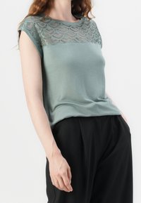 Světle zelený top s texturovanou krajkovou vrstvou na ramenou. Krátké rukávy a volný střih. Spárováno s černými, splývavými kalhotami.