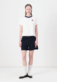 Hvid cropped t-shirt med en marineblå krave og lille logo. Marineblå shorts med logodetalje. Knælange hvide sokker og sorte flade sko.