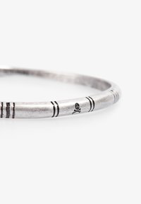 Pulsera de metal plateado con rayas grabadas y un pequeño motivo de calavera. Forma redonda con una textura suave y un acabado pulido.