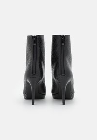 Paire de bottines noires à talons hauts avec fermetures éclair arrière et surface texturée à motifs, vues de dos sur fond blanc.