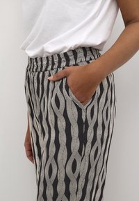 Un adulte portant un pantalon à motifs noir et blanc avec une main dans la poche, associé à une chemise blanche simple rentrée.