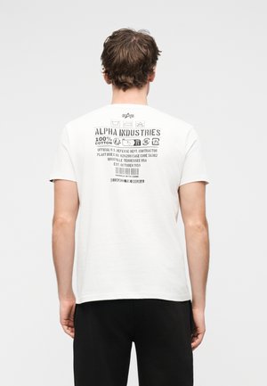 Homme vu de dos portant un t-shirt blanc Alpha Industries avec des textes et symboles noirs imprimés sur le haut du dos, associé à un pantalon noir.