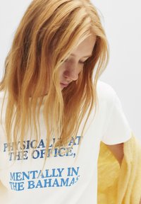 T-shirt en coton blanc avec un texte bleu indiquant : "PHYSIQUEMENT AU BUREAU, MENTALEMENT AUX BAHAMAS." Texture douce, manches courtes.
