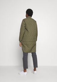 Chaqueta larga acolchada de color verde oliva con cuello alto, combinada con jeans grises y zapatillas blancas. La textura presenta un patrón de diamantes.