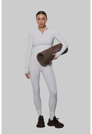 Kobieta w białym stroju sportowym trzymająca zwiniętą brązową matę do jogi z napisem „estio”, stojąca na jednolitym szarym tle.