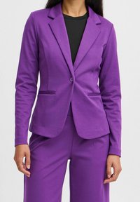 Kvinna som bär en figursydd lila blazer och matchande byxor över en svart topp, med minimal smyckeshållning på båda händerna.