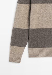 Rukáv hnědě a béžově pruhovaného svetru vyrobeného z měkké vlny, s žebrovanými manžetami a hladkou texturou. Klasický střih s horizontálními pruhy.