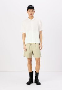 Light-colored, textured polo shirt with a collar, paired with beige shorts and black knee-high boots, standing against a white background.