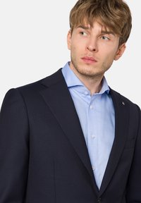 Granatowa marynarka z wyciętymi klapami, z pojedynczym zapięciem na guzik i teksturowanym materiałem; zestawiona z jasnoniebieską koszulą z guzikami pod spodem.