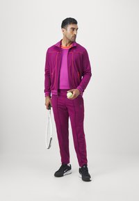Completo sportivo magenta con accenti arancioni. Include una giacca con zip e pantaloni aderenti. La persona tiene una pallina da tennis e una racchetta. Sneakers nere.