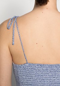 Top de tela texturizada azul y blanca con tirantes delgados y ajustables. Costura recogida visible a lo largo de la parte posterior. Tono de piel visible en la espalda.