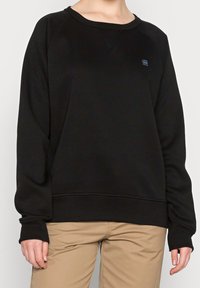 Persona che indossa un felpa nera a maniche lunghe con un piccolo logo sul petto e pantaloni beige, in piedi contro uno sfondo semplice.