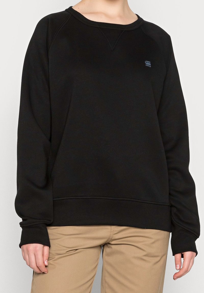 Persona che indossa un felpa nera a maniche lunghe con un piccolo logo sul petto e pantaloni beige, in piedi contro uno sfondo semplice.