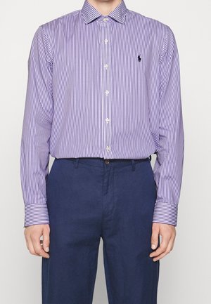 Mężczyzna ubrany w fioletowo-białą koszulę z pionowymi paskami i długim rękawem, wpuszczoną do ciemnoniebieskich spodni, stojący na jednolitym tle.