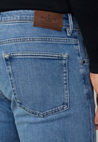 Vaqueros de denim azul ajustados con un parche de cuero marrón con el logo en la cintura y un bolsillo trasero con detalles de costura visibles.