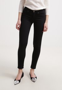 Černé skinny džíny s hladkou texturou a přiléhavým střihem, které mají přední zapínání na knoflík a pět kapsových stylů. V kombinaci s černými plochými botami.