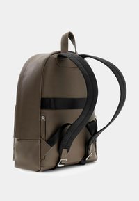 Mochila de tela marrón con una textura suave, que cuenta con una correa negra, un bolsillo con cremallera lateral y una asa en la parte superior para un transporte fácil.