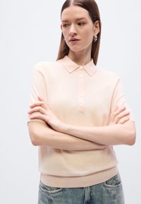 Πολό μπλουζάκι με κοντά μανίκια σε ανοιχτή ροδακινί απόχρωση, κατασκευασμένο από μαλακό πλεκτό ύφασμα, με ριμπ γιακά και έξι κουμπιά στο μπροστινό μέρος που ταιριάζουν χρωματικά.