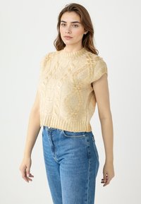 Crème-kleurige gebreide cropped trui met een hoge hals, korte mouwen, gestructureerde diamantpatronen en een geribde rand, gecombineerd met blauwe jeans.