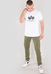 Vit kortärmad t-shirt med en svart grafisk logotyp, ihop med olivfärgade cargo-byxor med flera fickor som sitter åtsittande vid anklarna. Vita sneakers.