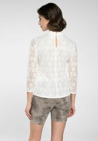 Weißes Spitzenoberteil mit Dreiviertelärmeln, ausgestattet mit einem Schlüsselverschluss hinten, kombiniert mit taupefarbenen Shorts. Texturierte und leichte Materialien.