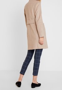 Beige uldfrakke med lige snit, detalje på bagsiden og påsyede lommer. Kombineret med marineblå ternede bukser og sorte slip-on sko.