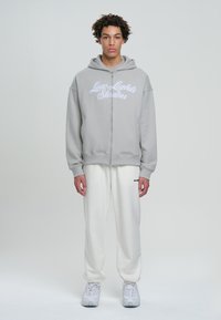Felpa grigia con zip e testo ricamato bianco, abbinata a pantaloni tuta color crema. Il modello è in piedi contro uno sfondo neutro, mettendo in mostra l'outfit.