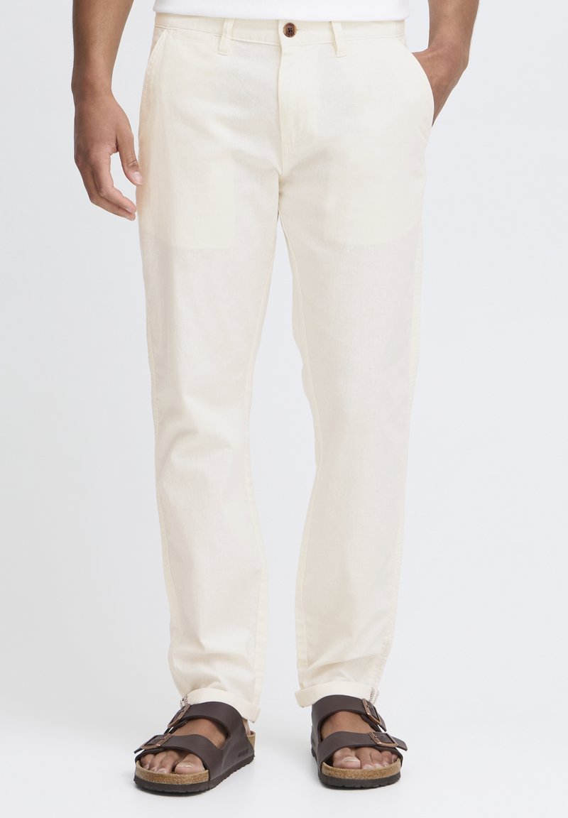 Partie inférieure du corps d'une personne portant un pantalon écru avec des poches avant et des sandales marron foncé à bout ouvert avec deux brides bouclées.