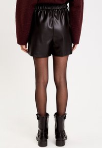 Shorts en simili cuir noir avec une taille élastique, associés à des bottines noires, sur un fond neutre. Collants translucides visibles.