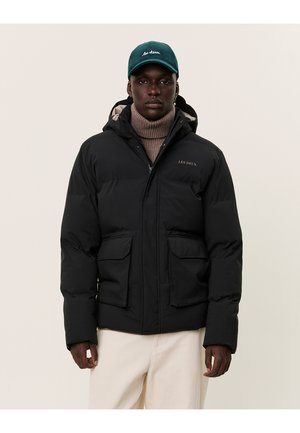 Black puffer jacket with a zip closure, two front pockets, and a hood. Worn over a brown turtleneck. Features an embroidered logo on the chest.