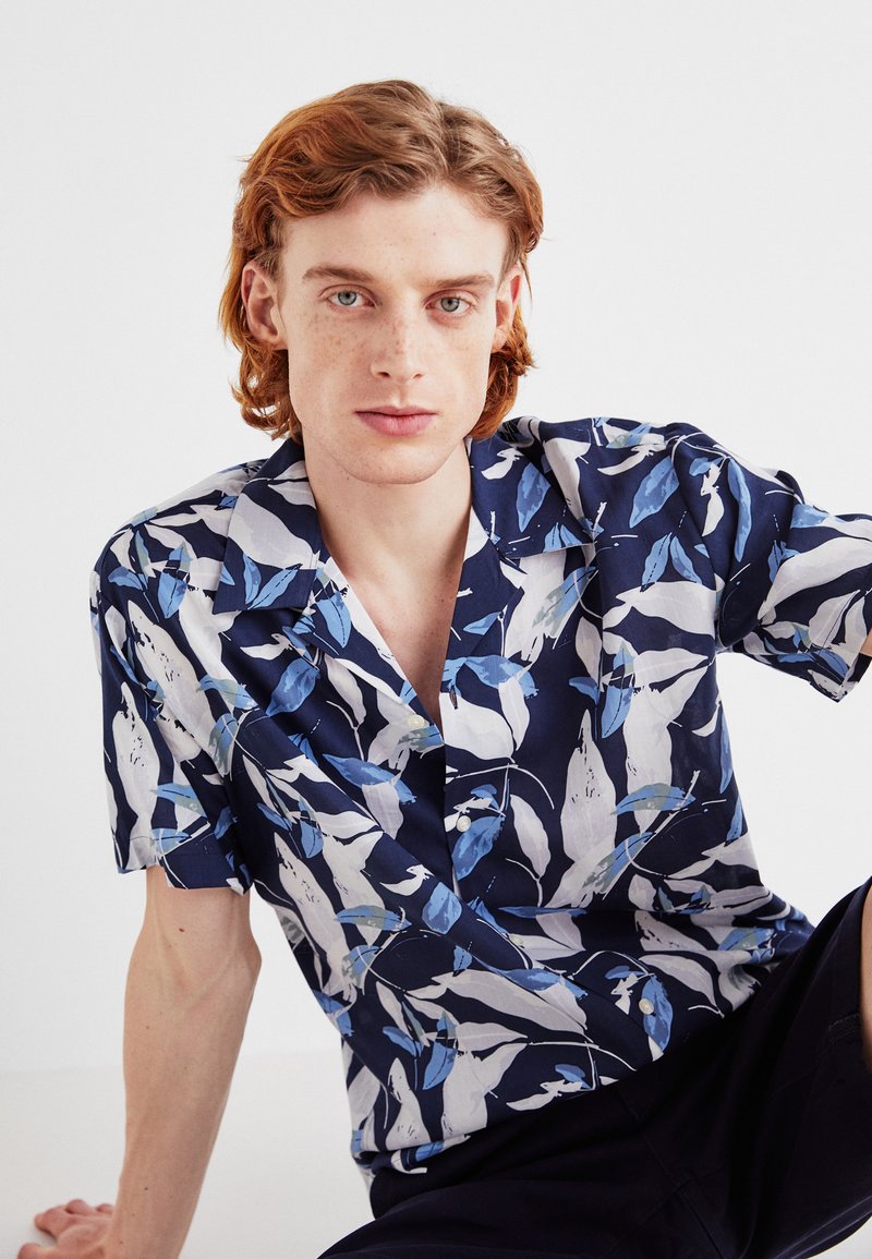 Camicia a maniche corte in navy con motivo floreale. Realizzata in tessuto leggero, presenta foglie bianche e azzurre, con colletto e bottoni.