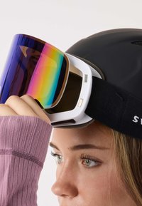 Gogle narciarskie z polaryzowanym, wielobarwnym szkłem i białą oprawą, umieszczone na czarnym kasku. Teksturę podkreśla dopasowany czarny pasek.