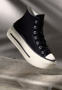 Svart høyhælte sneakers med glanset overflate, hvit gummisåle og kontrasterende svart stripe. Sølvøyer og snører fullfører designet.