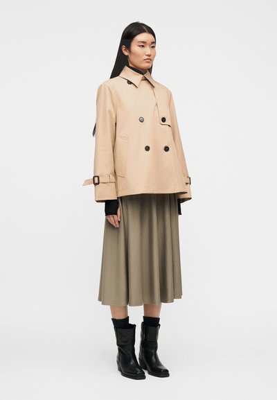 Γυναίκα με μπεζ σακάκι διπλής σειράς κουμπιών, ελιάς μίντι φούστα, μαύρες μπότες και μαύρο μακρυμάνικο τοπ από μέσα, στέκεται σε λευκό φόντο.