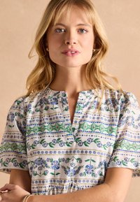 Šaty so vzorom kvetov z ľahkej tkaniny, s modrými a zelenými motívmi na bielom pozadí, s výstrihom v tvare lode a krátkymi rukávmi.