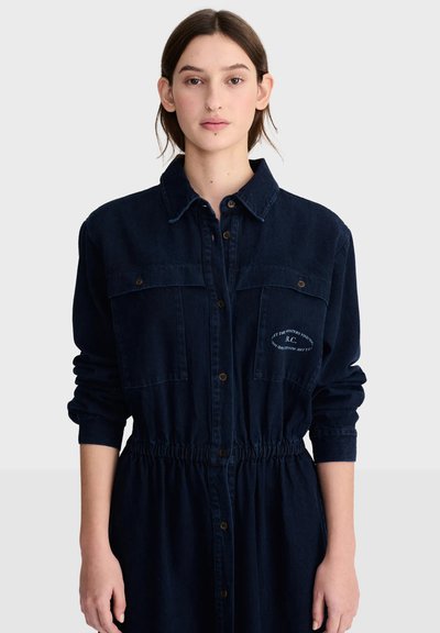 Jeune femme aux cheveux foncés porte une robe boutonnée bleu foncé à manches longues avec poches poitrine sur un fond clair uni.