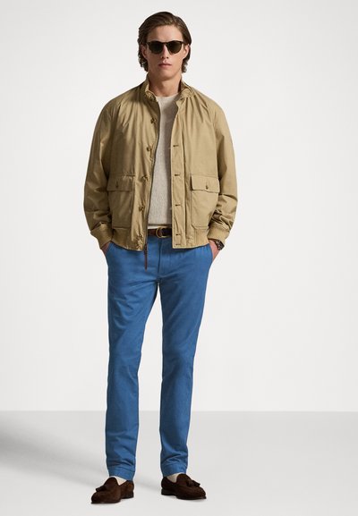 Casaco leve bege com fechos de botão, usado sobre um suéter claro, combinado com calças azuis e mocassins escuros. Óculos de sol completam o visual.