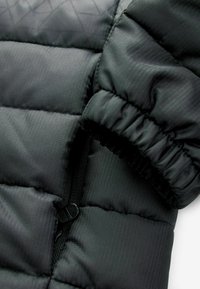 Tissu noir de veste en duvet avec un motif matelassé, doté d'une fermeture éclair sécurisée et d'un poignet élastique pour un ajustement près du corps. Surface texturée visible.