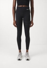 Schwarze aktive Leggings mit hohem Bund, die ein "STRONGER"-Logo tragen. Texturierte Streifen an den Seiten sorgen für Details. Kombiniert mit hellgrauen Schuhen.