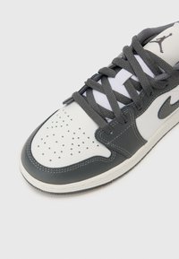 Baskets en cuir grises et blanches avec une boîte à orteils perforée, des lacets gris et un logo gris. Surface lisse avec des accents de surpiqûres contrastées.