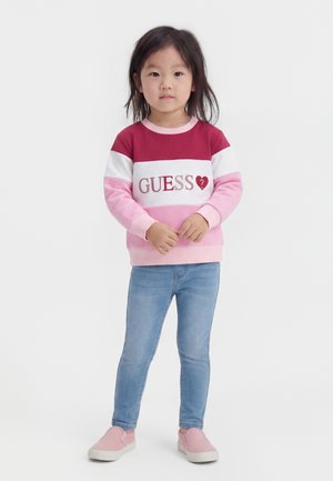 Ung pige står iført en rød, hvid og pink "GUESS" sweatshirt, blå jeans og pink slip-on sko mod en ensfarvet hvid baggrund.