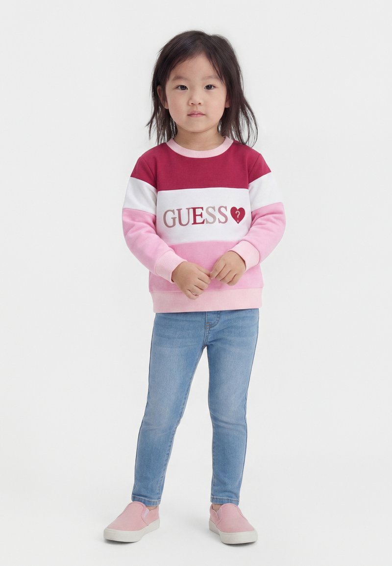 Jeune fille debout portant un sweat-shirt "GUESS" rouge, blanc et rose, un jean bleu et des chaussures roses sans lacets sur un fond blanc uni.