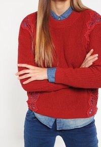Maglione rosso lavorato a maglia con scollo rotondo e dettagli in pizzo decorativi sui lati, indossato sopra una camicia in denim blu.