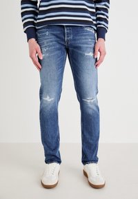 Jeans de mezclilla azul claro con corte Slim, que presentan áreas desgastadas y un lavado desteñido. Combinados con zapatillas blancas y una camisa a rayas en tono navy.