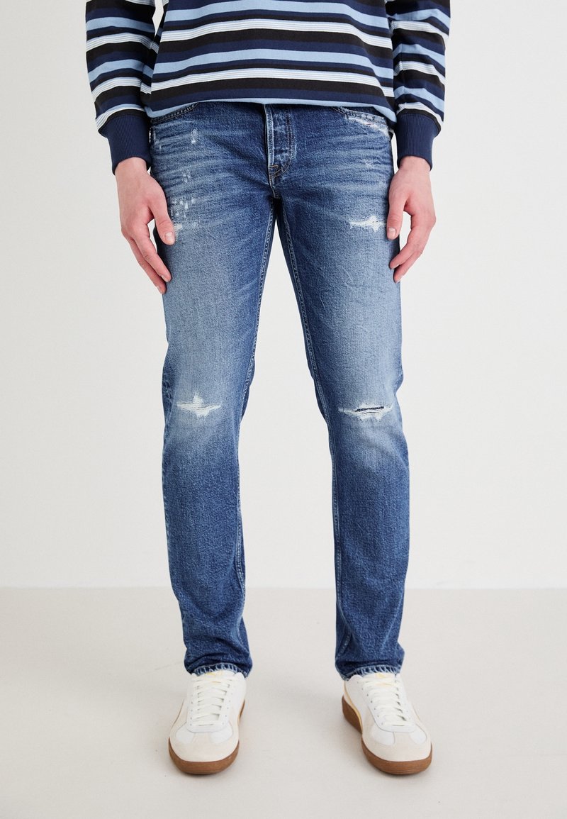 Jeans de mezclilla azul claro con corte Slim, que presentan áreas desgastadas y un lavado desteñido. Combinados con zapatillas blancas y una camisa a rayas en tono navy.